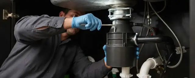Plumber mounting a garbage disposal under a kitchen sink with a screwdriver, trap and drain pipe in view